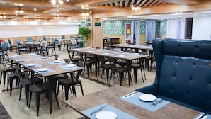 A dining area with multiple tables and chairs, a blue cushioned bench, and a ceiling with wooden beams and hanging lights.