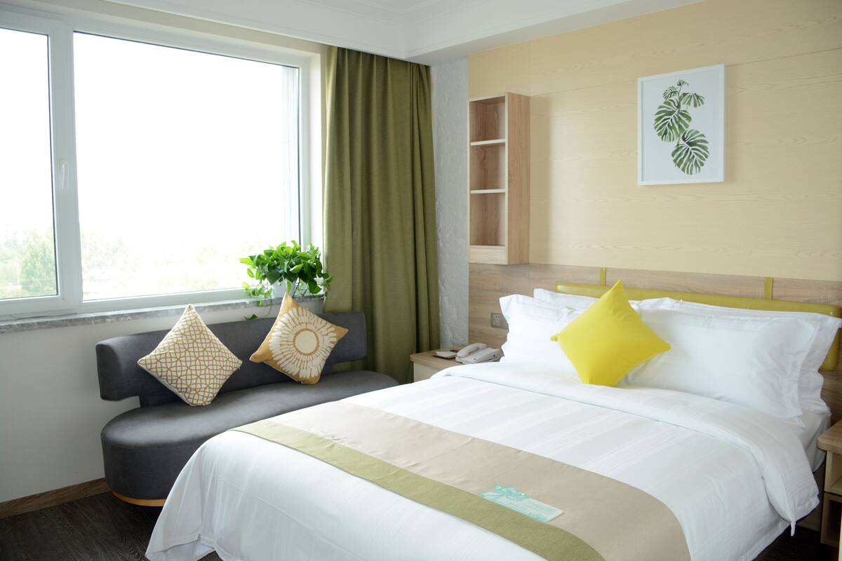 Een hotelkamer met een bed, een bank, een nachtkastje, een plant en een raam met gordijnen.