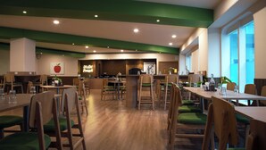 A spacious dining area with wooden tables and green cushioned chairs, a counter with a neon sign, and large windows allowing natural light. - Nest by TreeHouse Bhiwadi (Tijara)