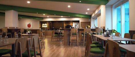 Un comedor espacioso con mesas de madera y sillas acolchadas verdes, una barra con letrero de neón y ventanas amplias que dejan entrar luz natural.