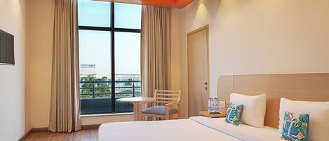 Quarto de hotel com uma cama grande, uma mesa pequena, uma cadeira e vista para a cidade através de uma janela ampla.