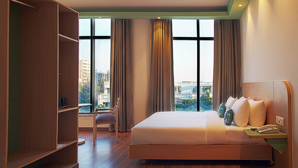 A hotel room with a large bed, a wardrobe, a chair, and a view of the city through large windows. - Nest by TreeHouse Bhiwadi (Tijara)