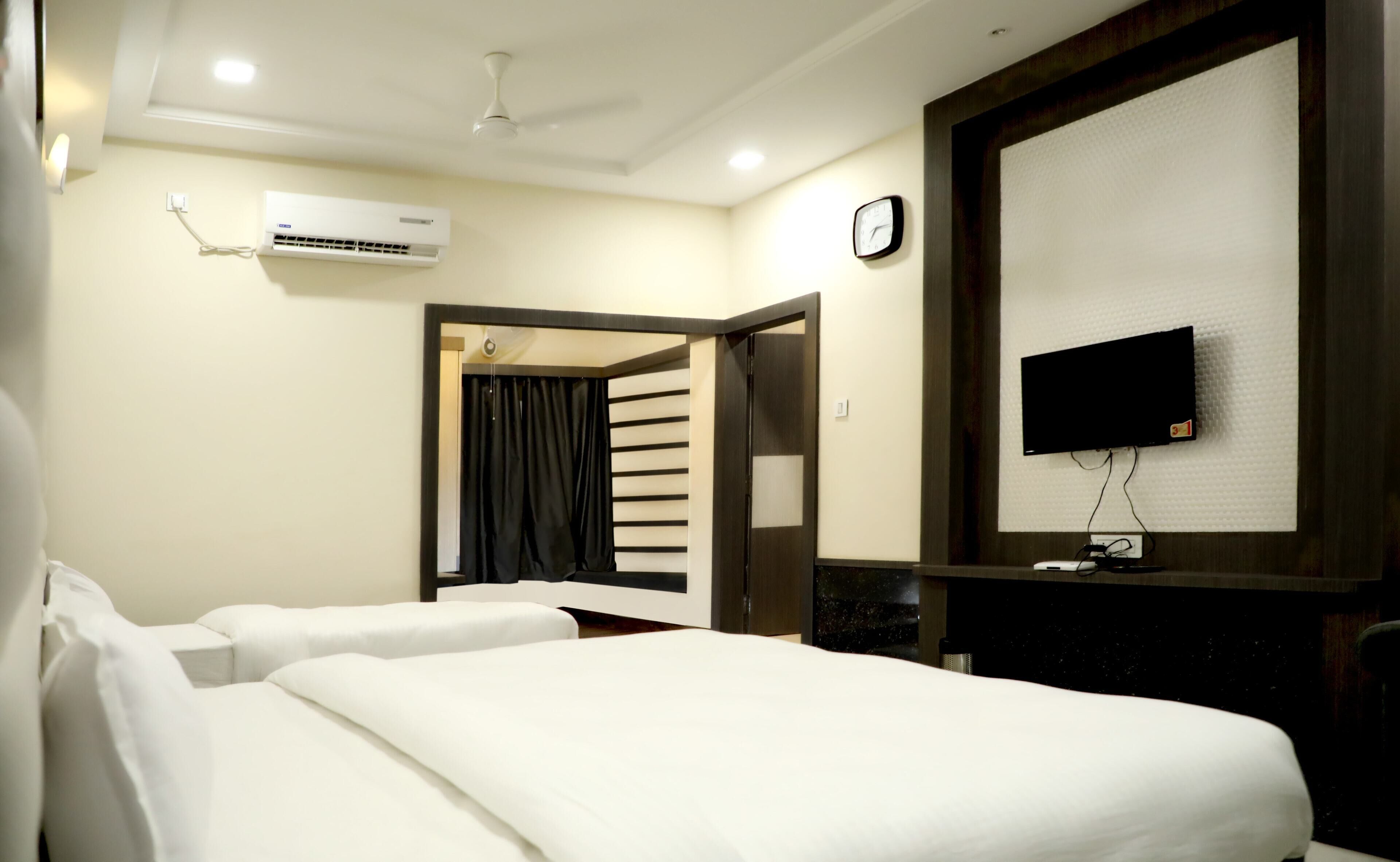 A hotel room with two beds, a flat-screen TV mounted on the wall, an air conditioning unit, a clock, and a door leading to another room.