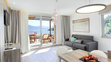 Un salón moderno con un sofá gris, una mesa de comedor de madera con fruta, y vistas al mar a través de grandes ventanales.