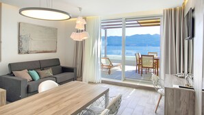 A modern hotel room with a sofa, dining table, and chairs, a TV, and a large window with a view of the sea and mountains. - Kalamota Beach House (Dubrovnik)