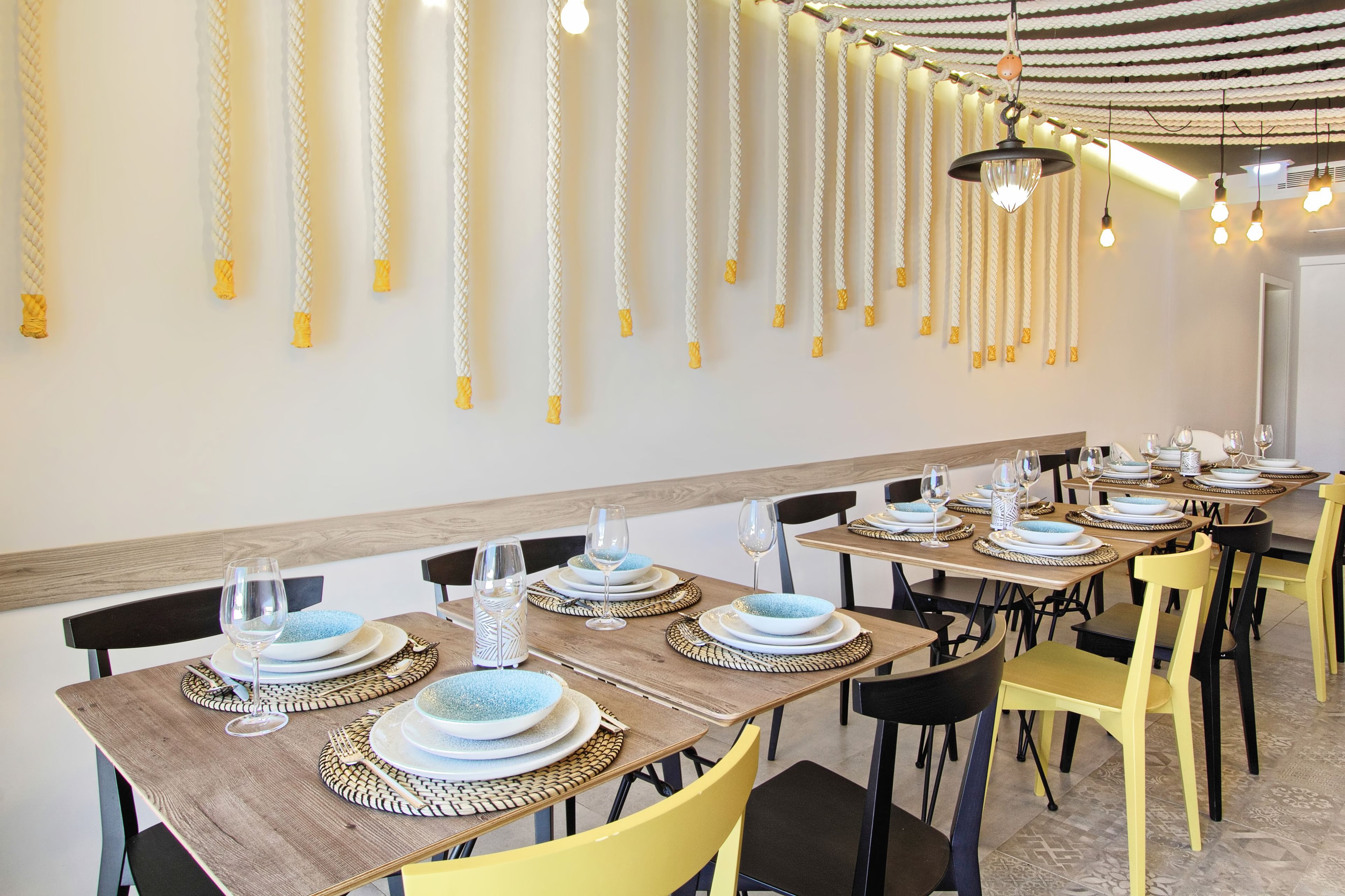 Un restaurante con mesas de madera y sillas amarillas, con platos azules y tazones blancos, y luces colgantes en el techo.