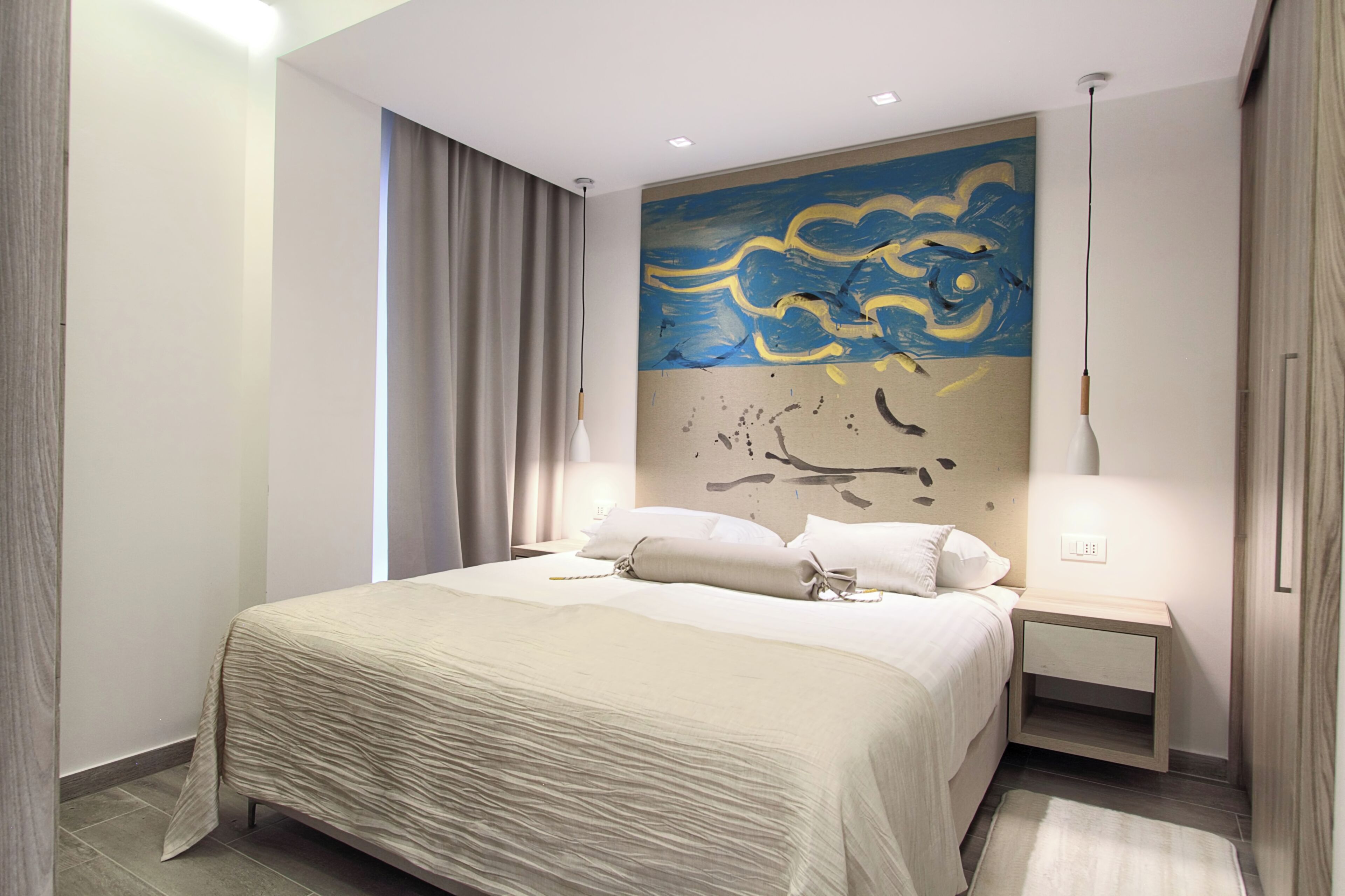 Una habitación de hotel moderna con una cama grande, una mesita de noche, un cuadro en la pared y una ventana con cortinas.