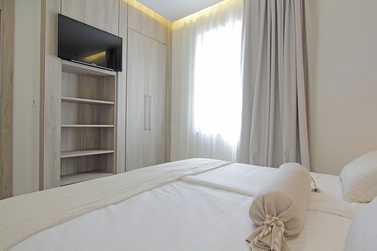 Un dormitorio con cama, televisor montado en un armario de madera y ventana con cortinas.