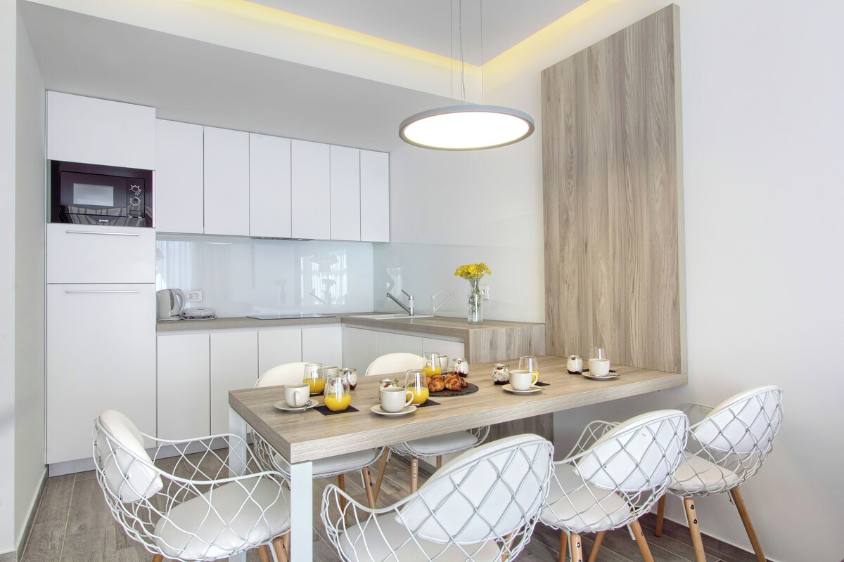 Una cocina moderna con armarios blancos, encimera de madera y una zona de desayuno con tazas, un tostador y un florero con flores.