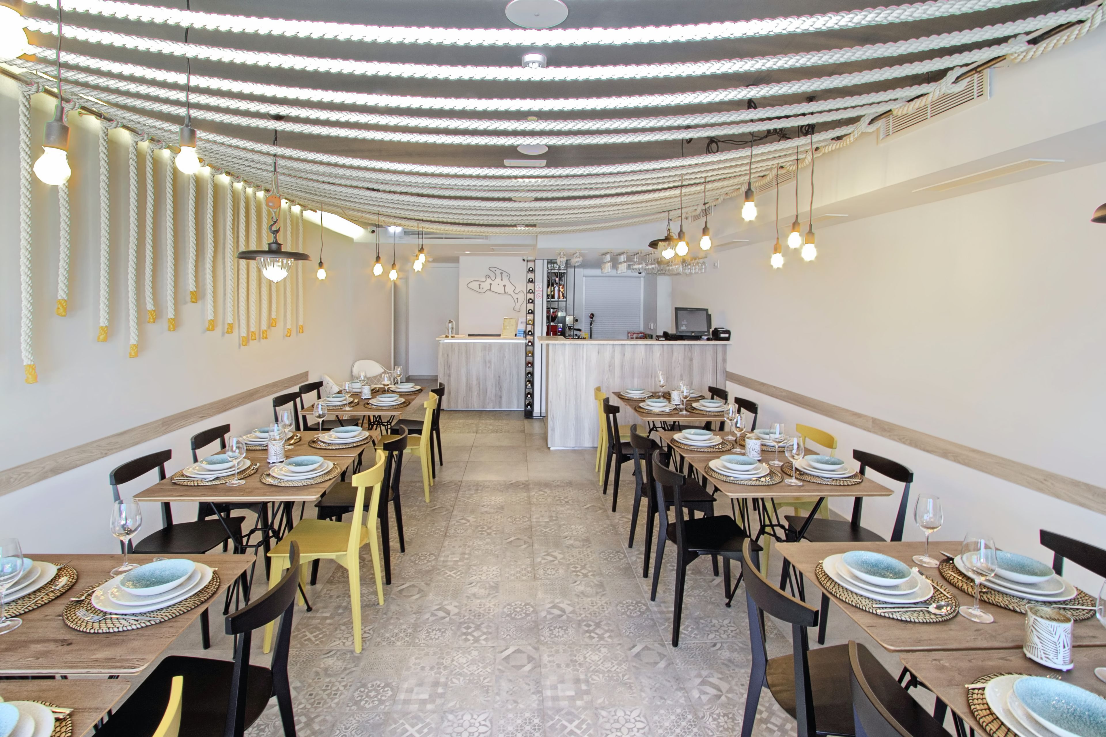 A dining area with multiple tables set for guests, featuring a patterned floor and a ceiling with hanging light fixtures.
