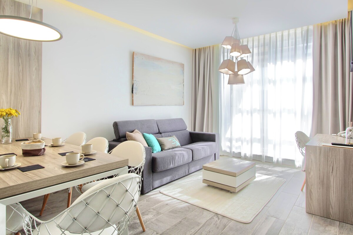 A modern living room with a grey sofa, a dining table with chairs, a coffee table, and a large window with curtains.