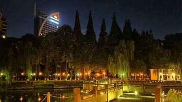 A nighttime view of a park with a bridge, trees, and a building with illuminated signage in the background.