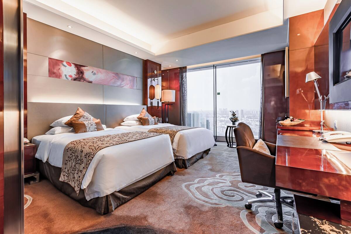 A modern hotel room with two beds, a desk, and a large window offering a city view.