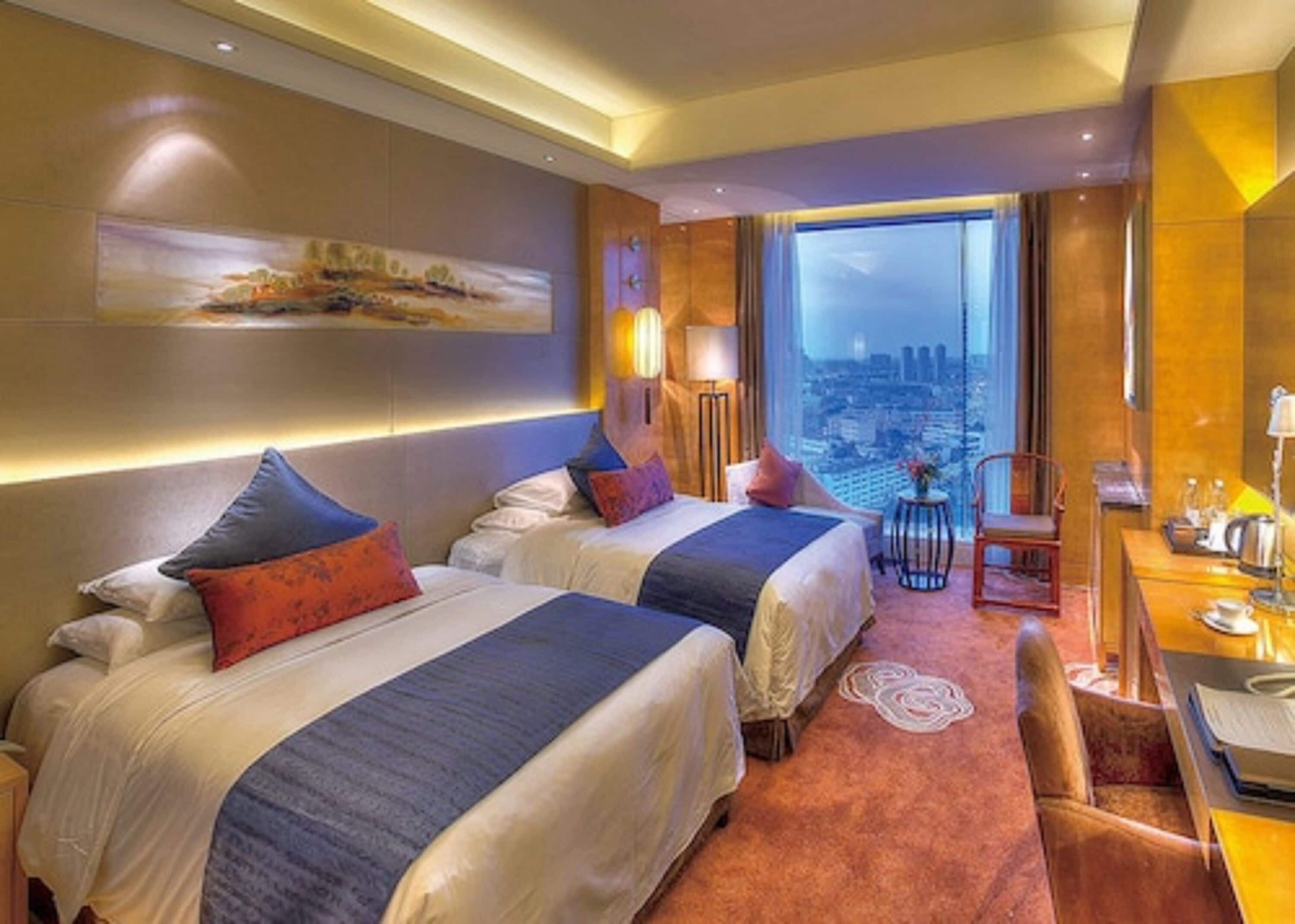 Habitación de hotel con dos camas, un escritorio, una silla y un amplio ventanal con vistas a la ciudad.