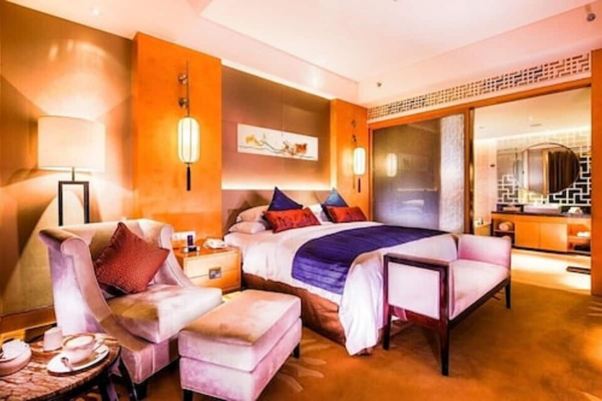 A modern hotel room with a large bed, a sitting area with a chair and ottoman, a bedside table with a lamp, and a large mirror.