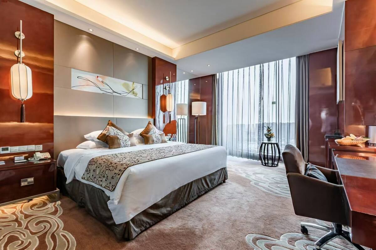 A modern hotel room with a large bed, a desk, and a view of the city through large windows.