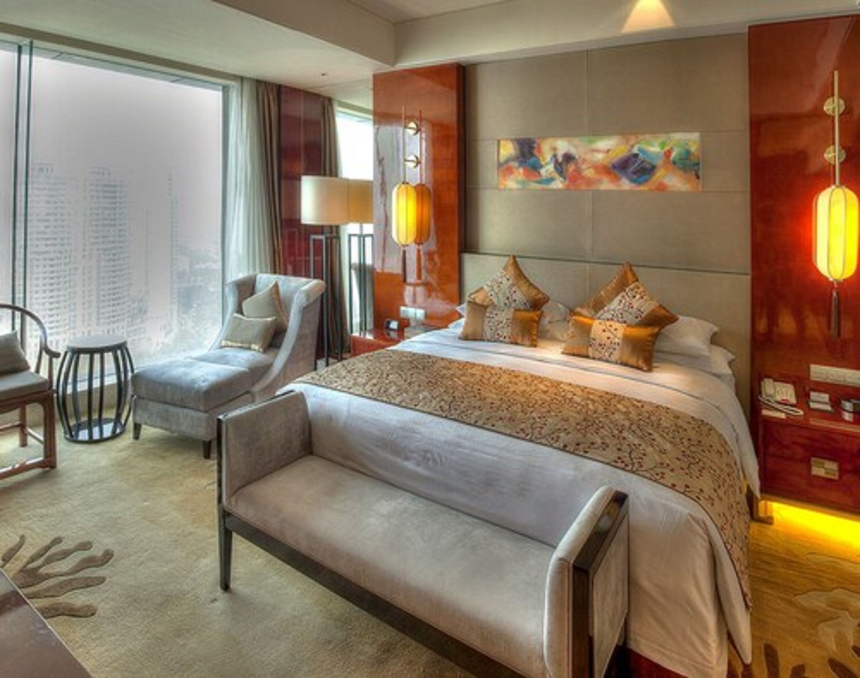 A hotel room with a large bed, a sofa, a chair, and a desk. There is a window with a view of buildings.