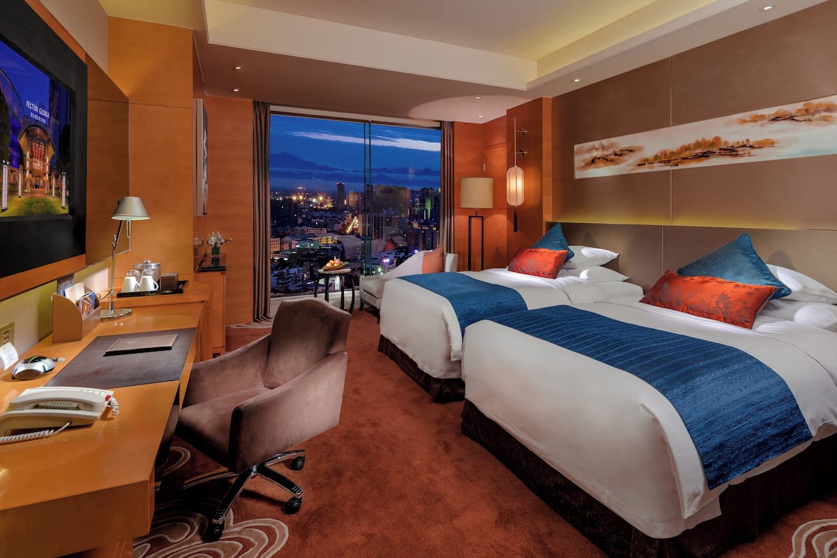 A hotel room with two beds, a desk, a chair, and a large window with a city view.