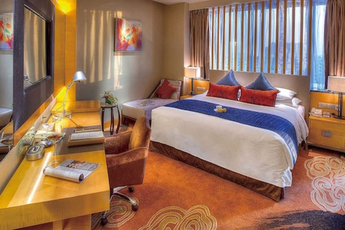 A hotel room with a large bed, a desk with a lamp, a chair, and a view of the city through the window.