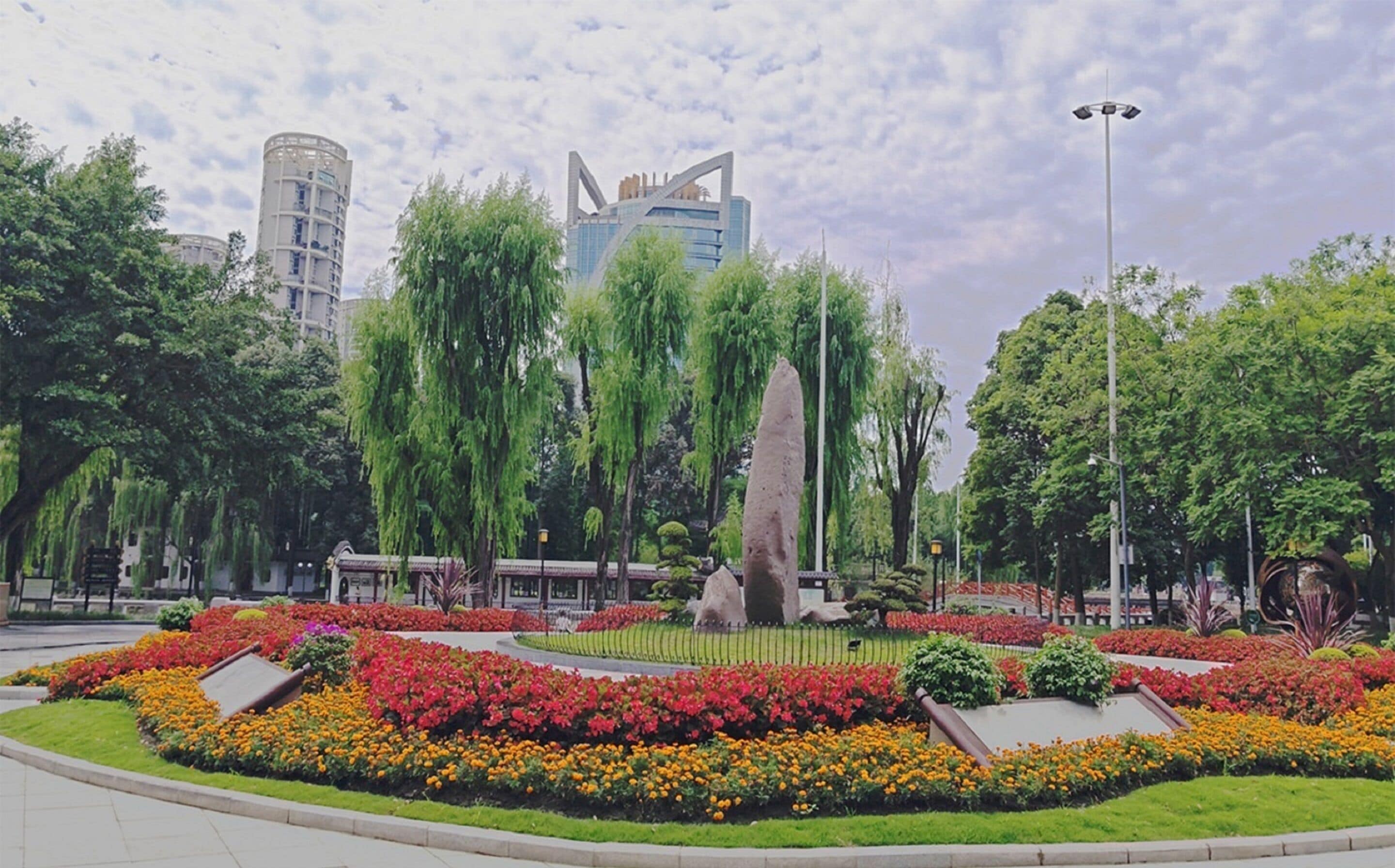 A public park with a central monument, surrounded by colorful flower beds and tall trees.