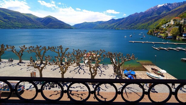 A lakeside view with mountains, a dock with boats, and a pedestrian walkway with trees. - Hotel Elvezia Ascona (Ascona)
