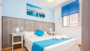 Siististi järjestetty hotellihuone, jossa on sänky, yöpöytä, seinälle ripustettu taulu ja ikkuna, jossa on sininen sälekaihdin.