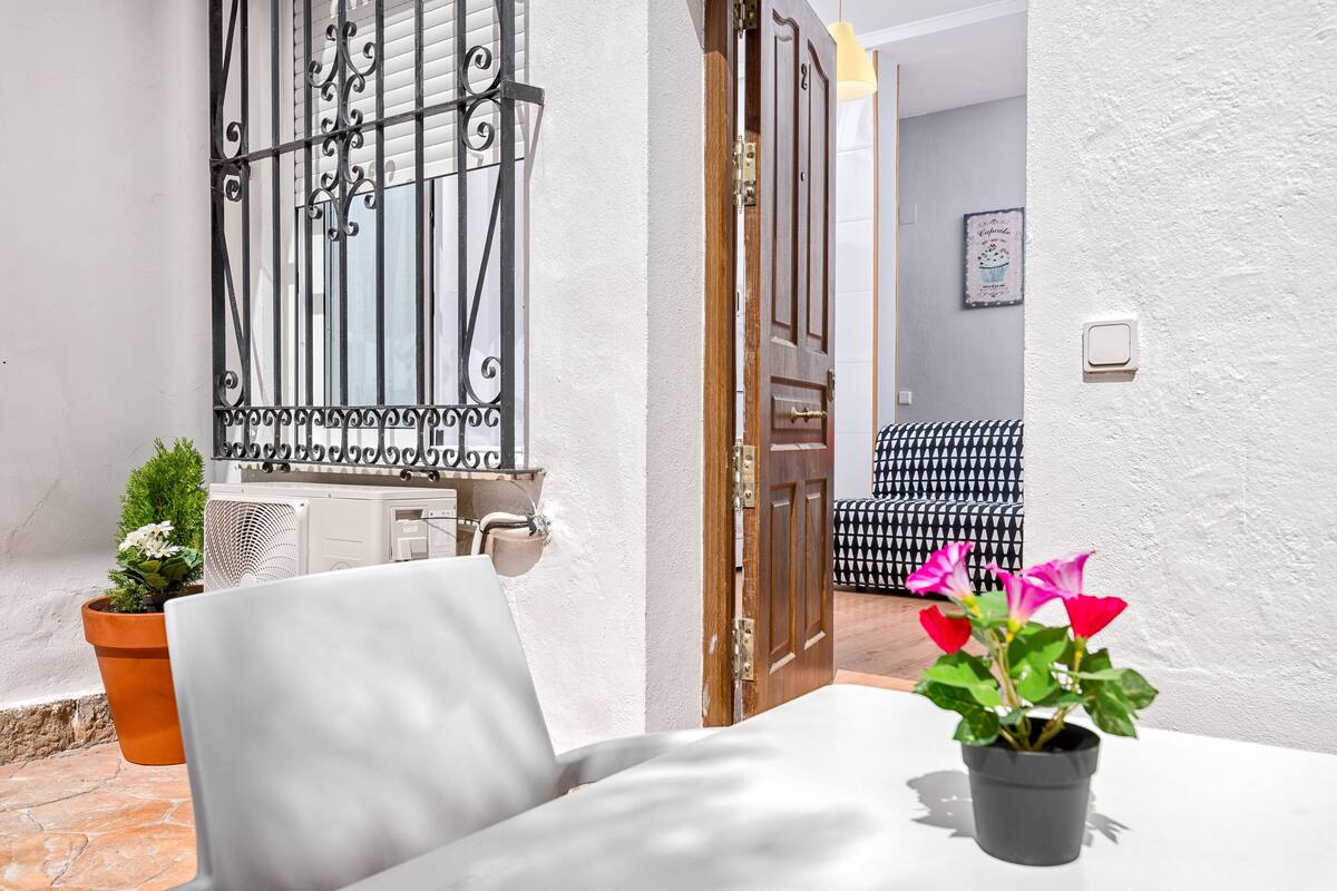 A white chair with a potted plant, a wooden door, and a patterned sofa in a well-lit room with a white wall and a window with black iron bars.