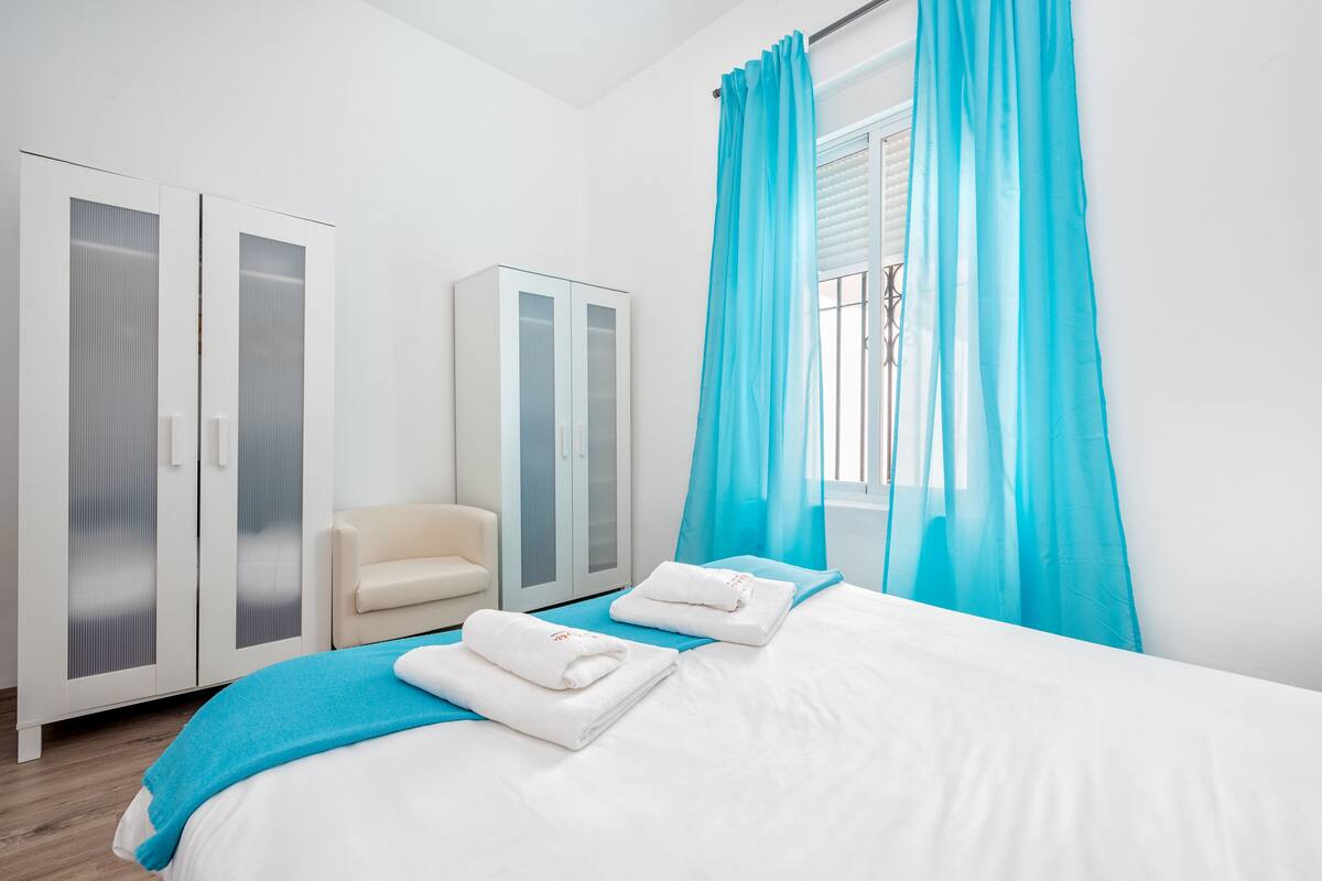 A hotel room with a bed, two white wardrobes, a beige chair, and a window with blue curtains.