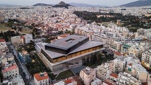 Edifici modern amb teulada plana i grans finestres, envoltat d'una zona urbana densa amb diversos edificis i una muntanya al fons.