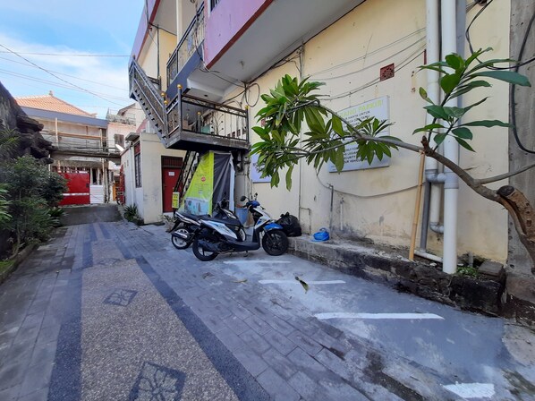 Een smalle straat met een geparkeerde scooter en motor, een rood hekwerk, en een kleine boom.
