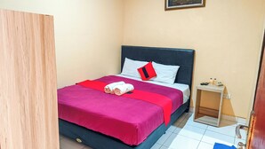 Een net opgemaakt bed met een rood en zwart kussen, een klein nachtkastje met een fles en een telefoon, en een schilderij aan de muur.