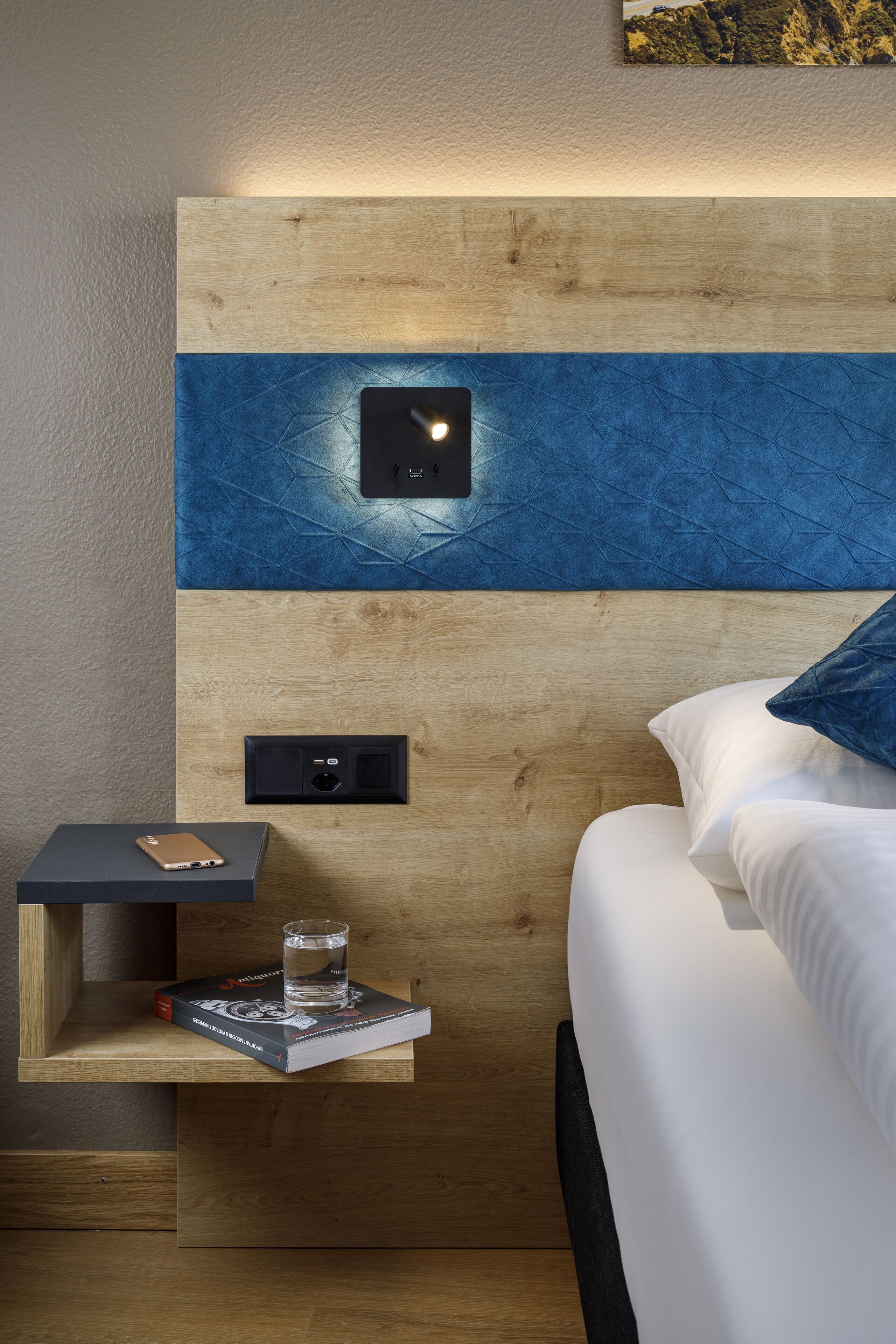 A modern hotel room with a wooden headboard, a bedside table with a book and a glass, and a power outlet.