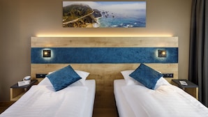 A hotel room with two beds, a large picture of a coastal landscape, and a wooden headboard with integrated lighting. - Hotel California (Zürich)