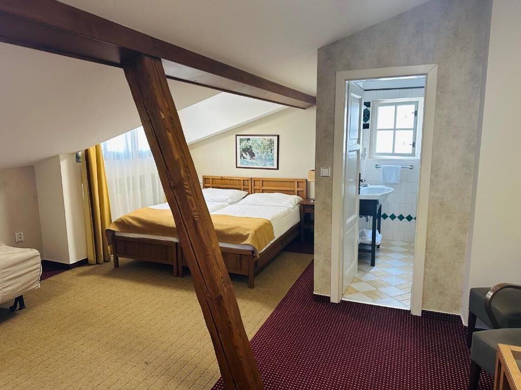 Un dormitorio con cama, una viga de techo de madera, alfombra roja y un baño visible a través de una puerta abierta.