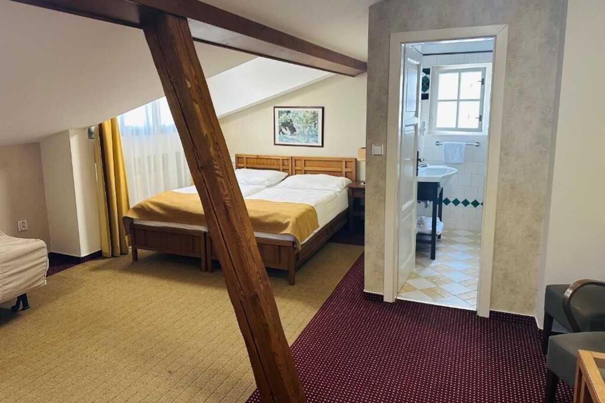 A bedroom with a bed, a wooden ceiling beam, a red carpet, and a bathroom visible through an open door.