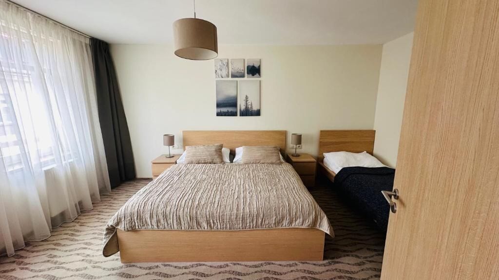 A hotel room with a large bed, two bedside tables, a hanging lamp, and a view of the city through sheer curtains.