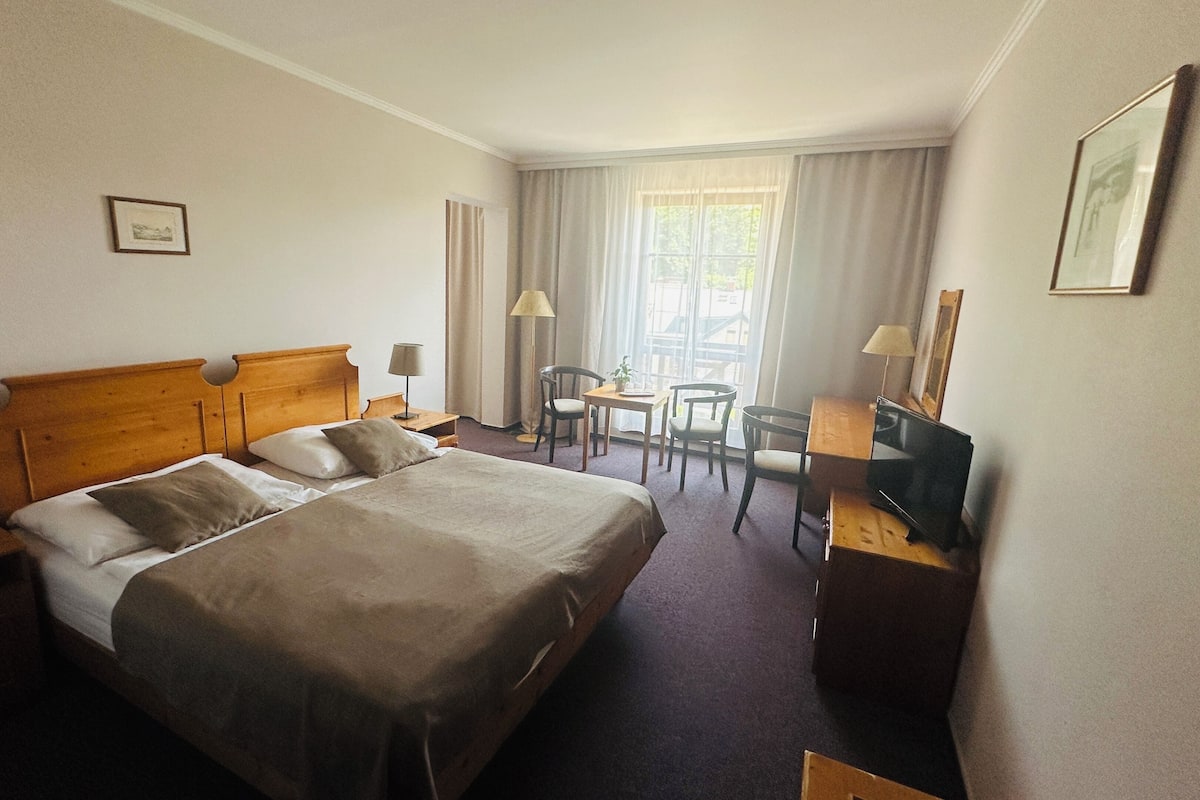 A hotel room with a large bed, a desk with a chair, a small table with two chairs, a lamp, and a window with curtains.