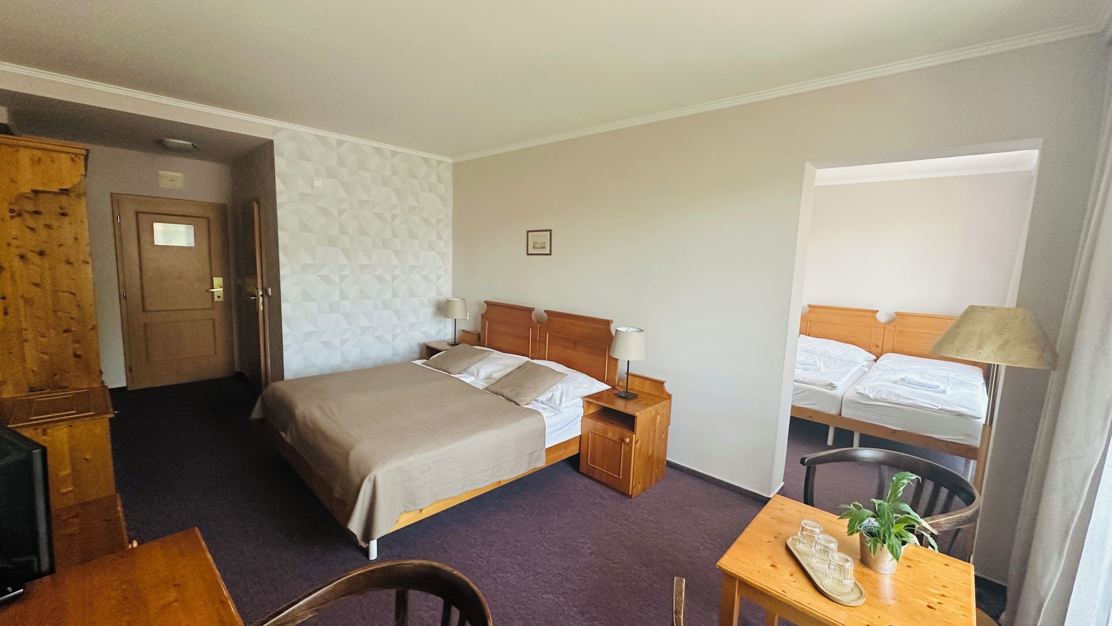 A hotel room with a bed, bedside tables, a desk, a chair, a small table with a plant, and a window.