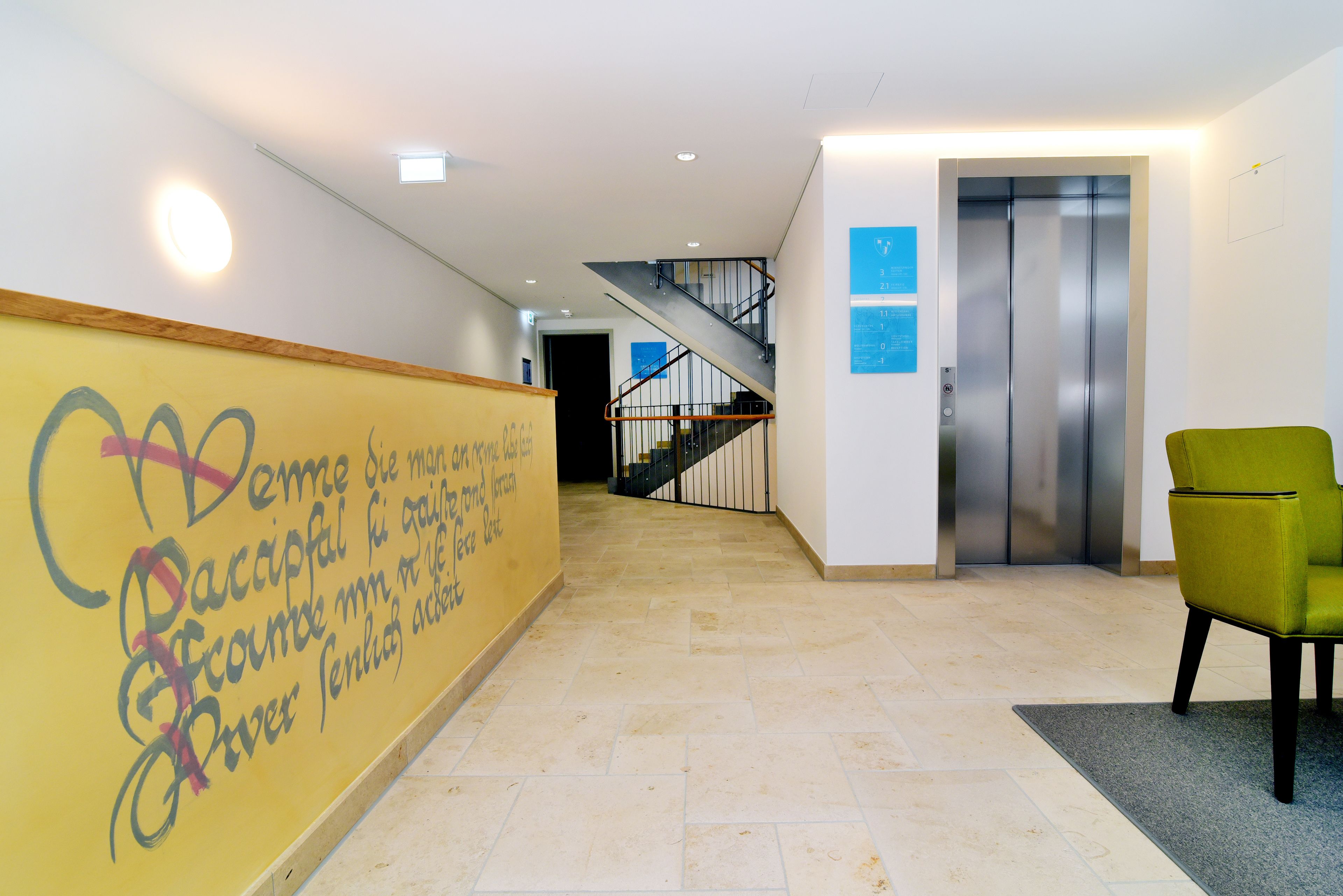 A hallway with a yellow wall featuring graffiti, a green chair, and an elevator.