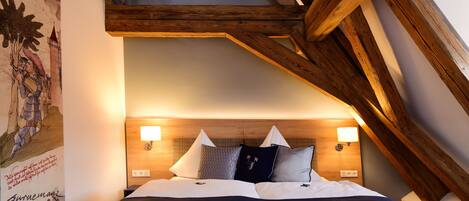 Un dormitorio con techo inclinado, vigas de madera, una cama con muchos cojines e iluminación en la pared.