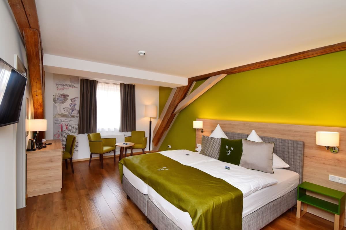 A hotel room with a bed, two chairs, a desk, a TV, and a window with curtains.