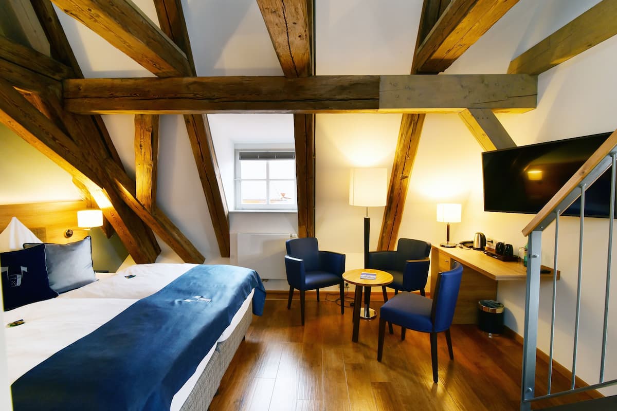 A room with a bed, two chairs, a desk, a TV, and wooden beams.