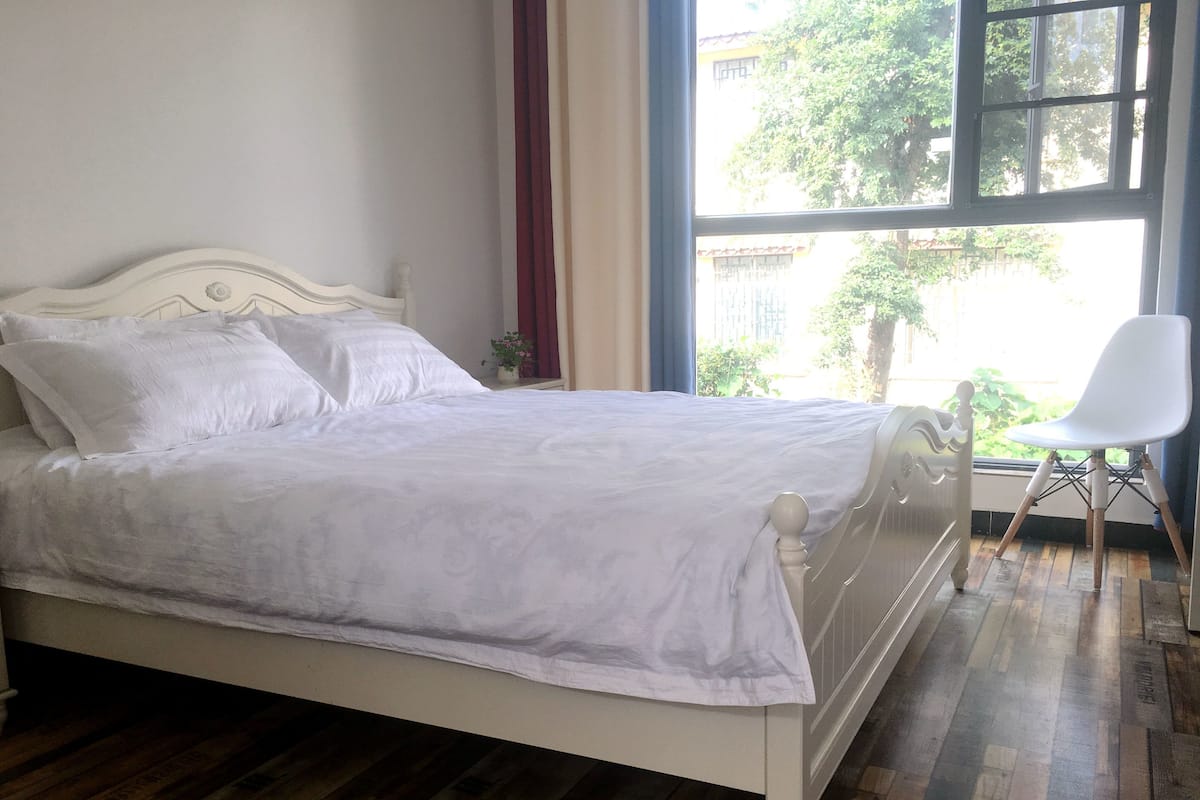 Una cama bien hecha con ropa de cama blanca en una habitación con suelo de madera y un ventanal con vistas al exterior.