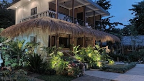 Ein modernes Haus mit einem Strohdach, umgeben von üppigen tropischen Pflanzen und einem gepflegten Garten.