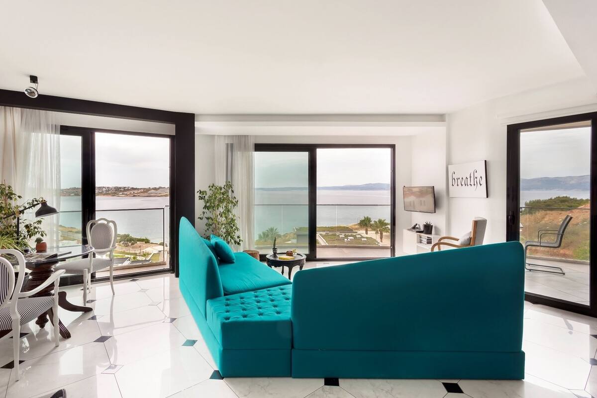 Ein modernes Wohnzimmer mit einem türkisfarbenen Sofa, einem Essbereich mit Tisch und Stühlen und großen Fenstern mit Blick auf die Landschaft.