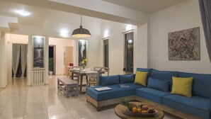 A modern living room with a blue sofa, a wooden coffee table with fruit, and a dining area with a table and chairs. - Villa Iasmi (Chania)