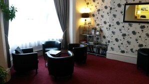Habitación de hotel con alfombra roja, sillas de cuero negro, una mesita, una estantería y una lámpara.