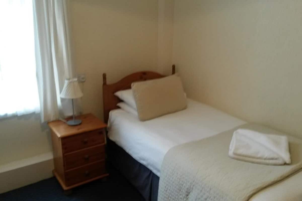 A small, neatly made bed with a wooden headboard, a bedside table with a lamp, and a window with curtains.