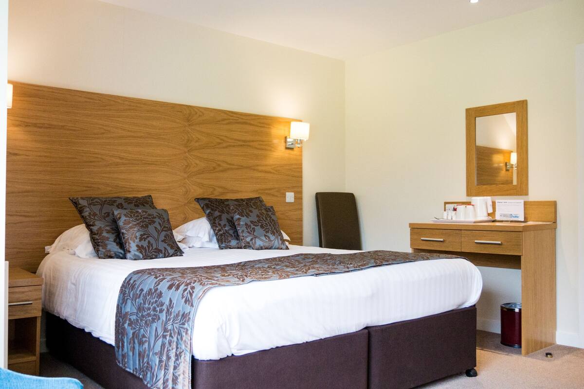 A hotel room with a large bed, a wooden headboard, a bedside table, a desk with a mirror, and a trash can.