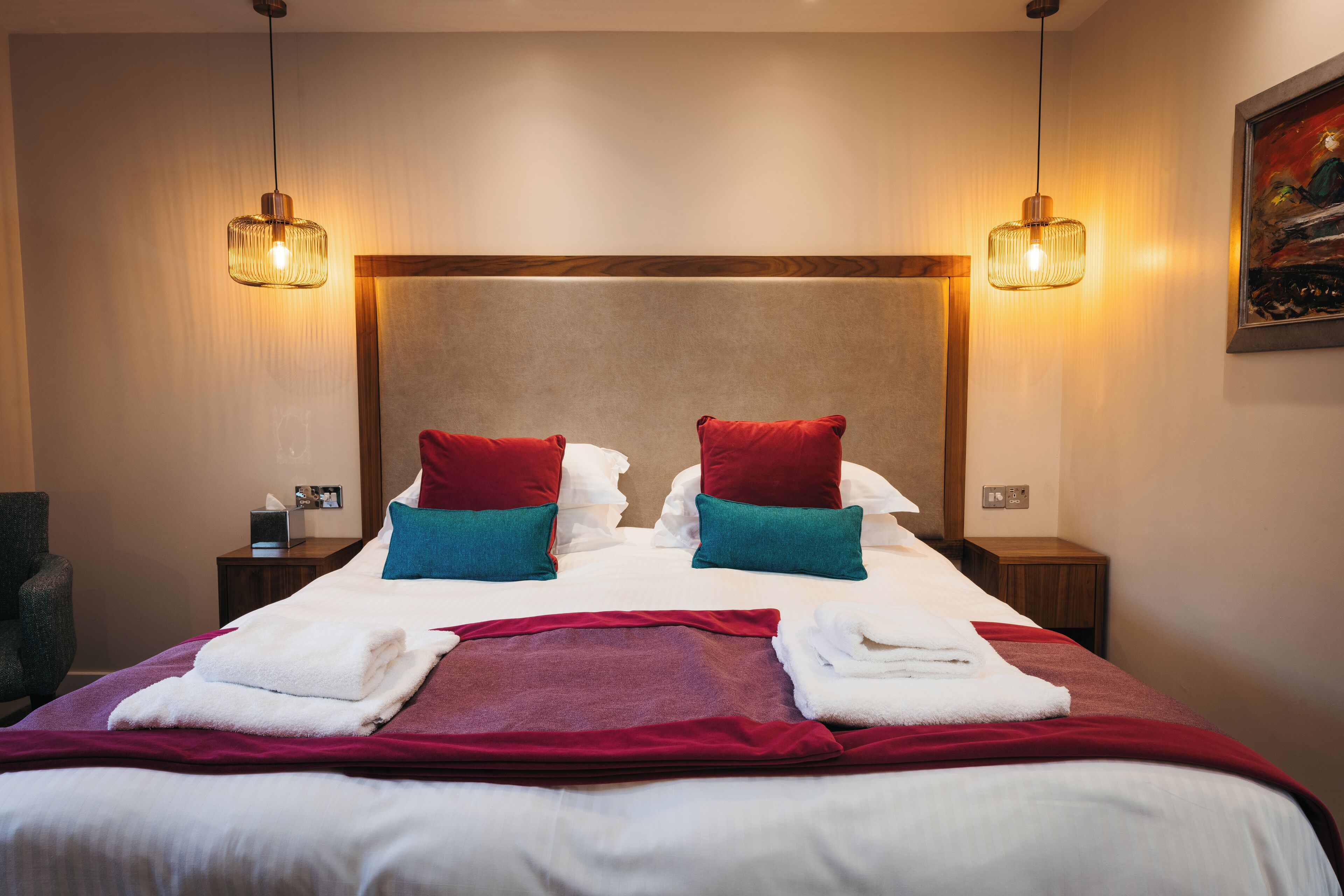 A hotel room with a large bed, two red and one teal pillow, a red throw blanket, and two bedside lamps.